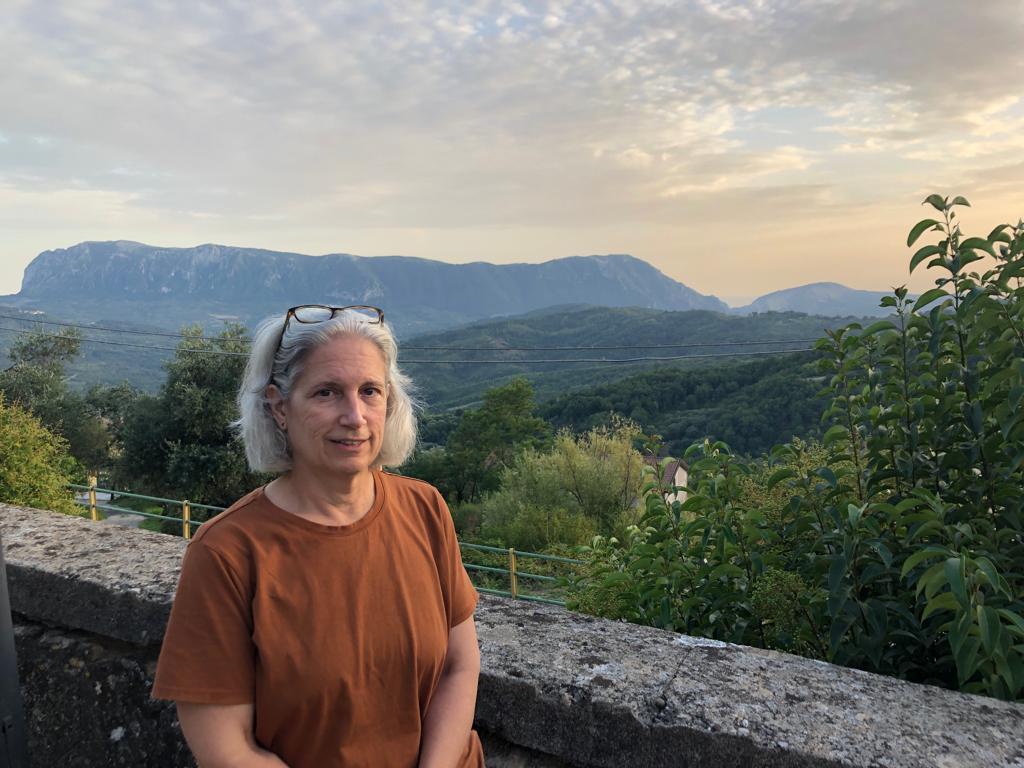 Photograph of woman with white hair wearing a brown shirt, standing outdoors in front of a low stone wall, overlooking mountains and a green valley in the background