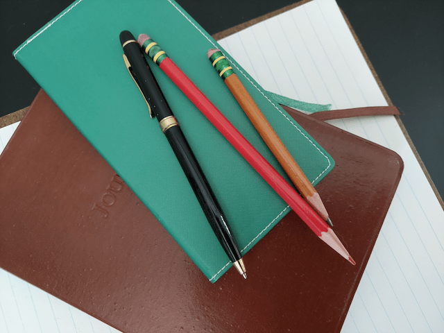 A black pen, red pencil, and brown pencil laid on a small green journal, which is on a brown leather journal, with ruled paper in the background